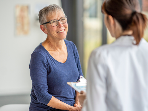 Elderly woman smiles while discussing her health with a doctor holding a clipboard in clinic