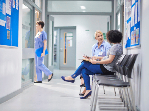 Two women talk in a clinic waiting area while a nurse walks down the hallway